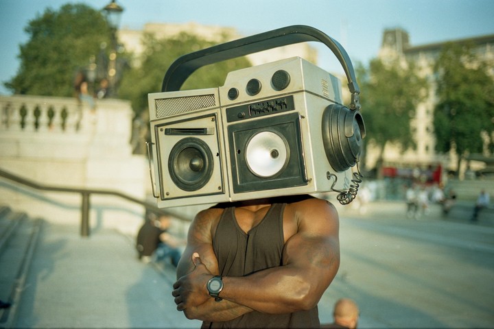 A man with a boombox on his head stands with his arms crossed on a sunny street.