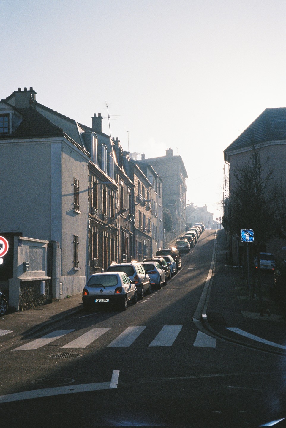 A street with parked cars on the left and buildings on both sides stretches uphill into a hazy sunrise.