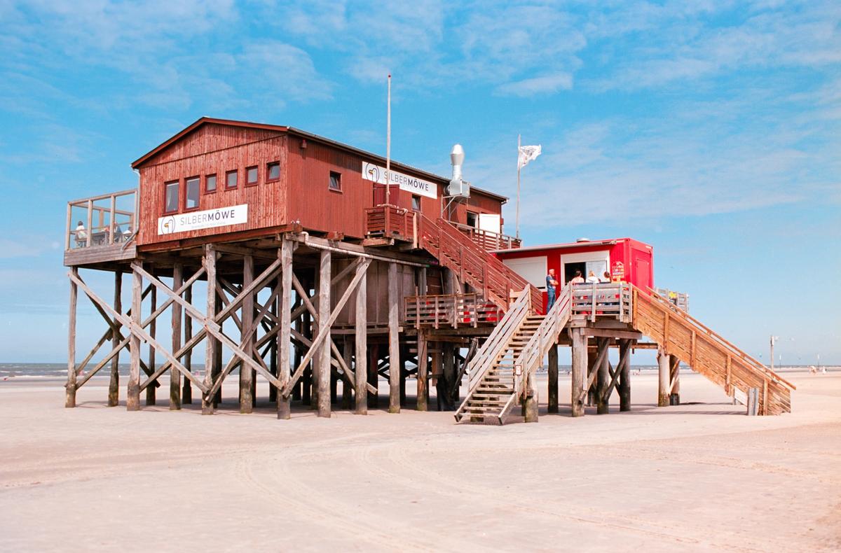 A red stilt house with wooden stairs stands on a sandy beach under a blue sky.