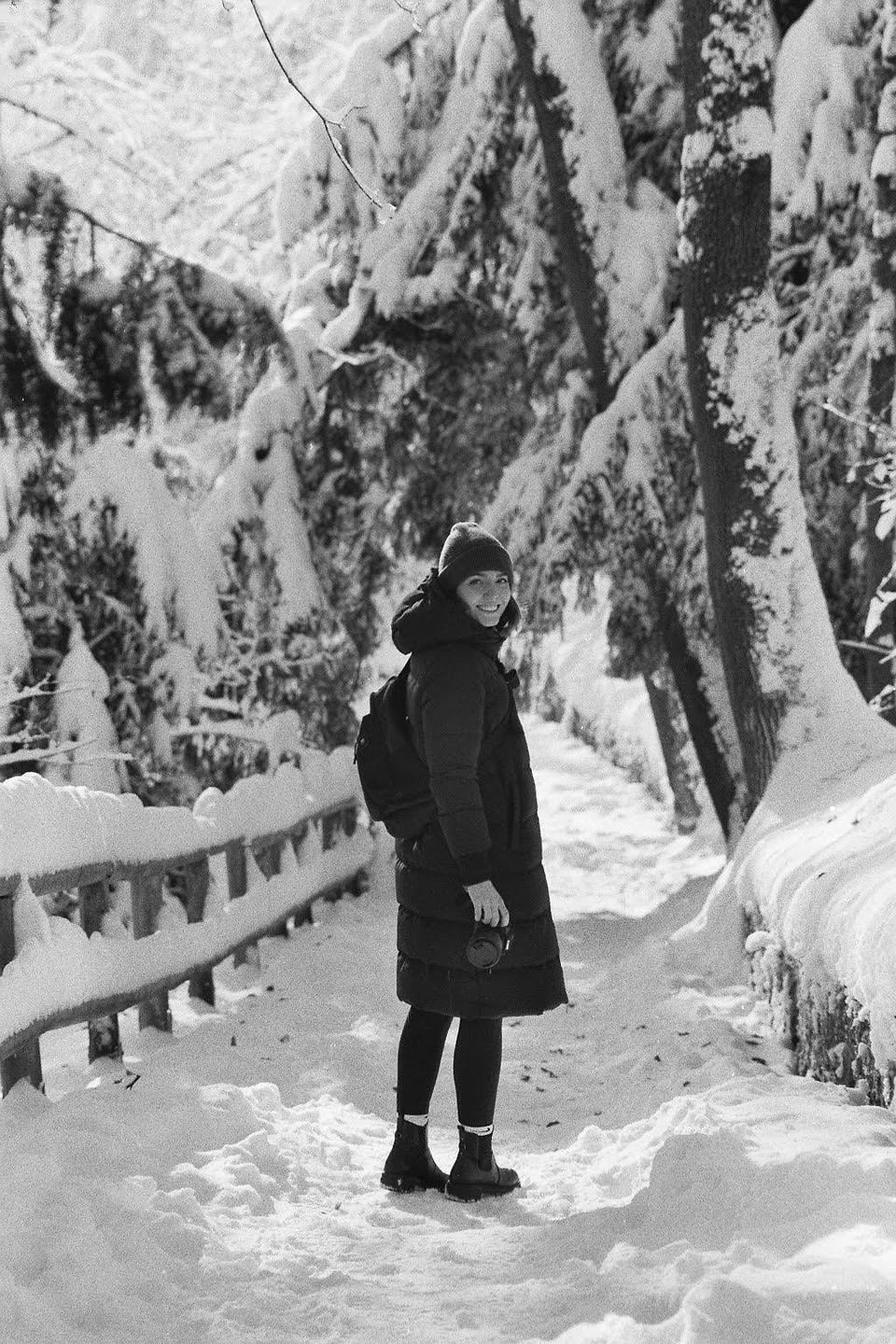 A woman wearing a winter coat and hat stands on a snowy path next to a snow-covered fence and trees.