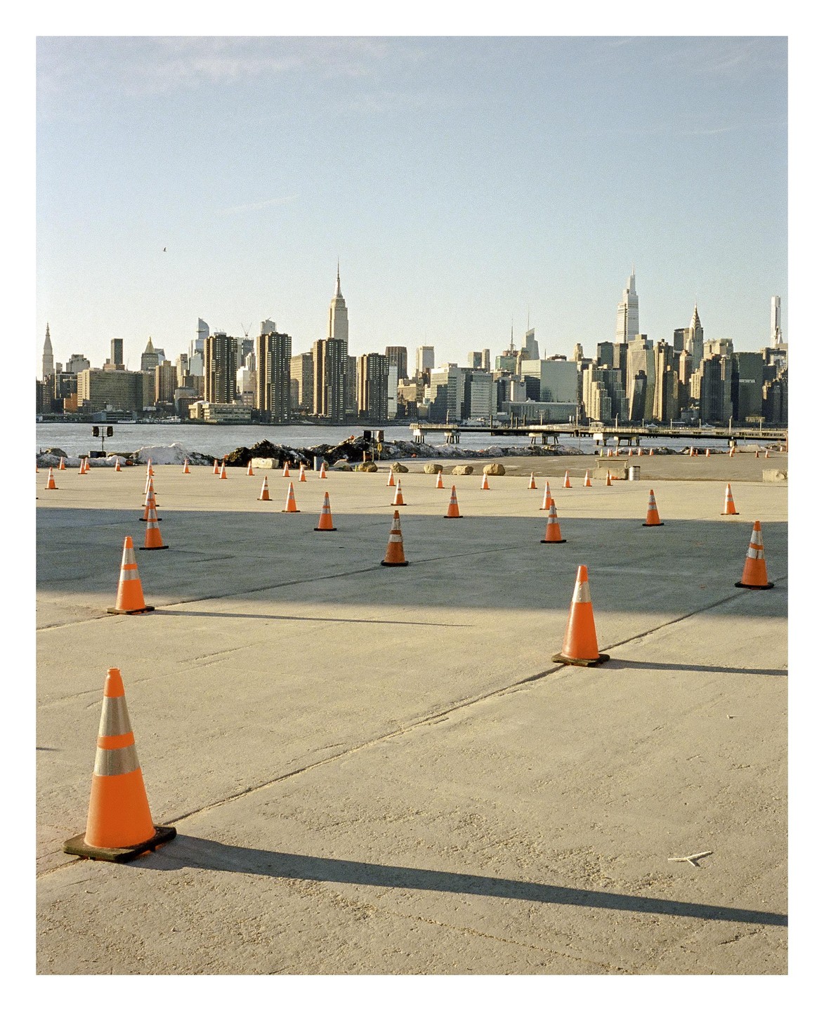 Orange traffic cones are scattered across a concrete lot with a city skyline and river in the background.