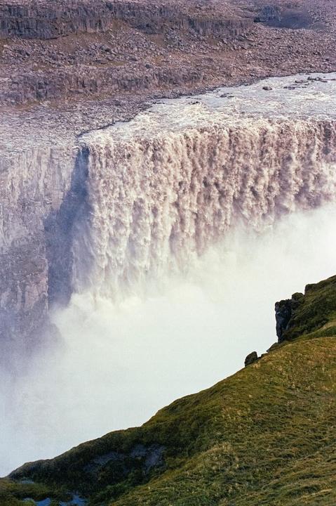 A powerful waterfall cascades over a rocky cliff, creating a misty cloud below, with a grassy hill in the foreground.