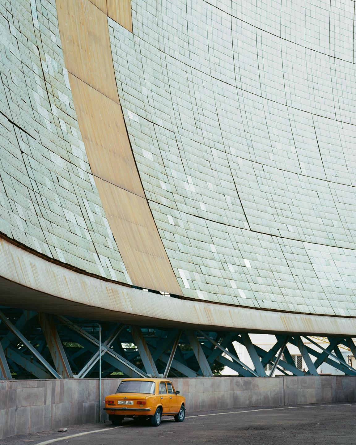 An orange car is parked next to a large, curved building with a textured facade.