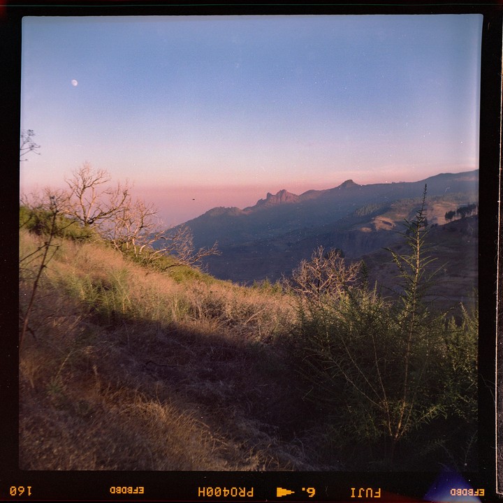 A moon is visible in the sky above a mountain landscape at dusk.