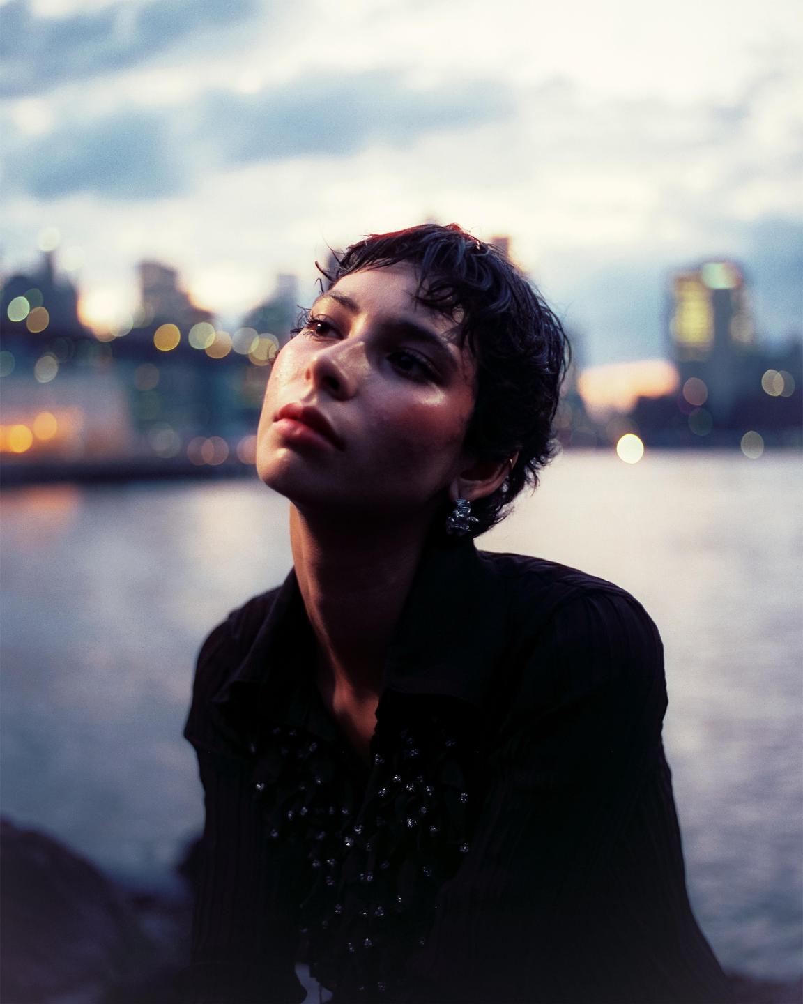 A woman with short, dark hair looks up with a city skyline and water in the background at dusk.