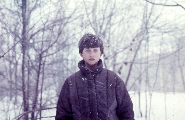 A boy with snow in his hair and on his jacket stands in a snowy forest.