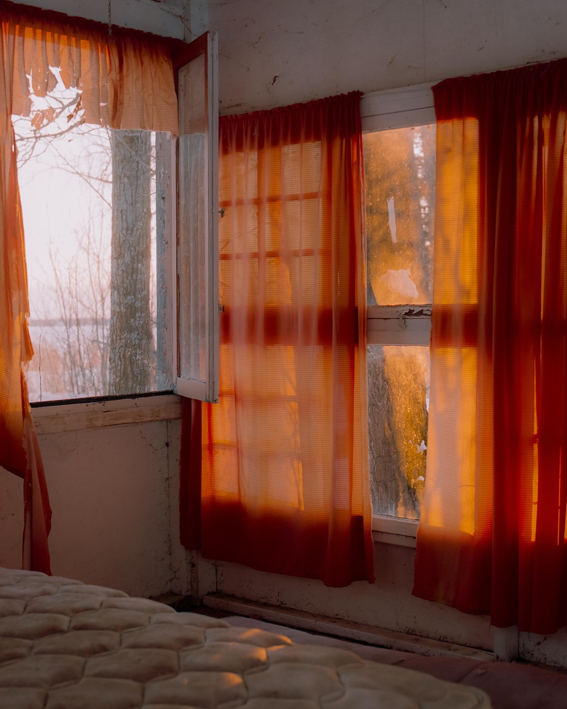Sunlight streams through red curtains and an open window into a room with a mattress in the foreground.