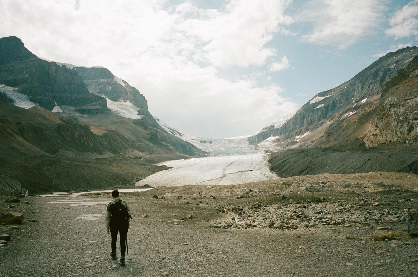 A person with a backpack walks on a rocky path towards a large glacier surrounded by mountains.