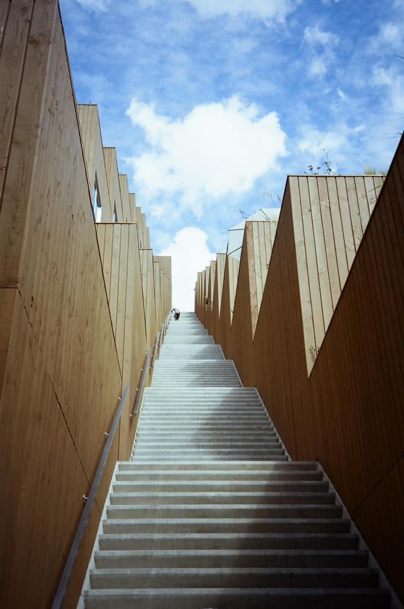 A long, narrow concrete staircase with wooden walls on either side leads up to a bright blue sky with white clouds.
