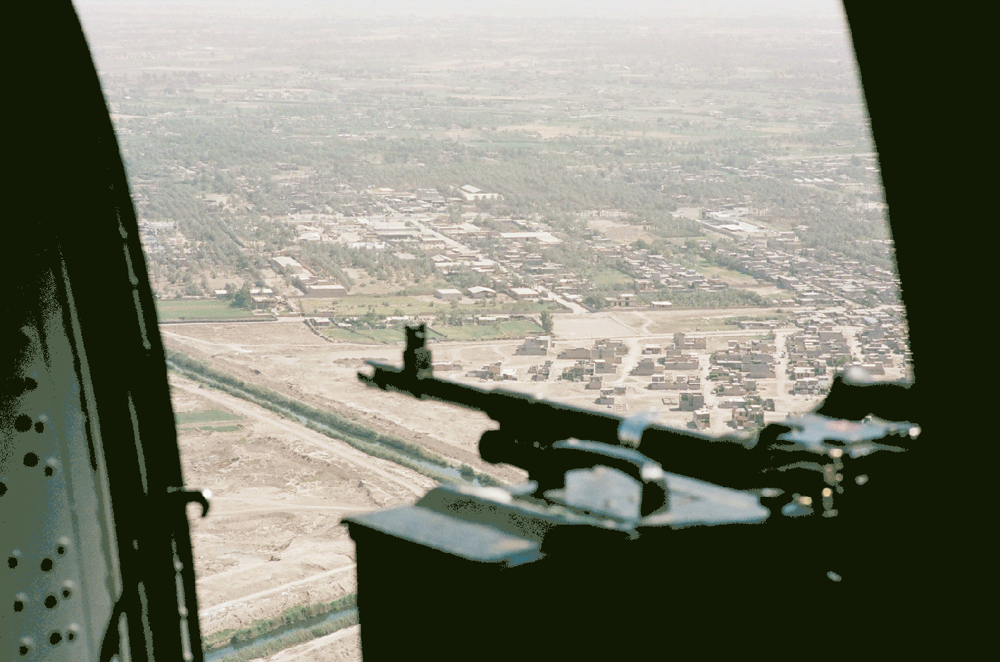 A machine gun is visible in the foreground with a town and fields in the background.