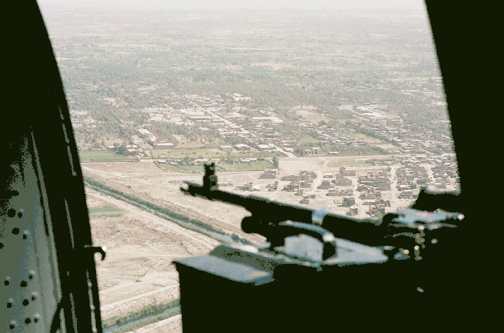 A machine gun is visible in the foreground with a town and fields in the background.