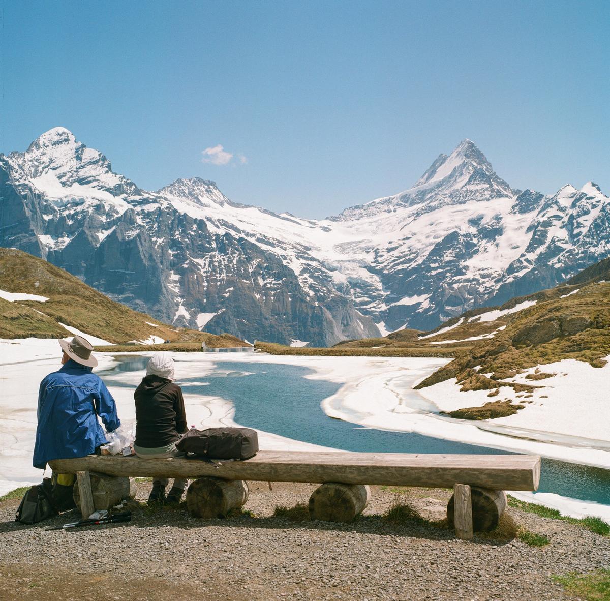 Two people sit on a wooden bench overlooking a partially frozen lake with snow-capped mountains in the background.