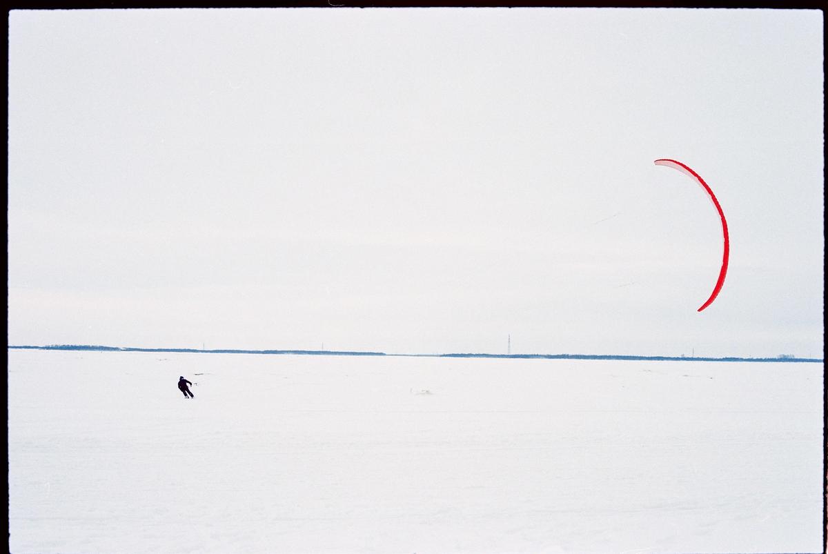 A person on skis is pulled by a red kite across a vast, snow-covered landscape under a pale sky.
