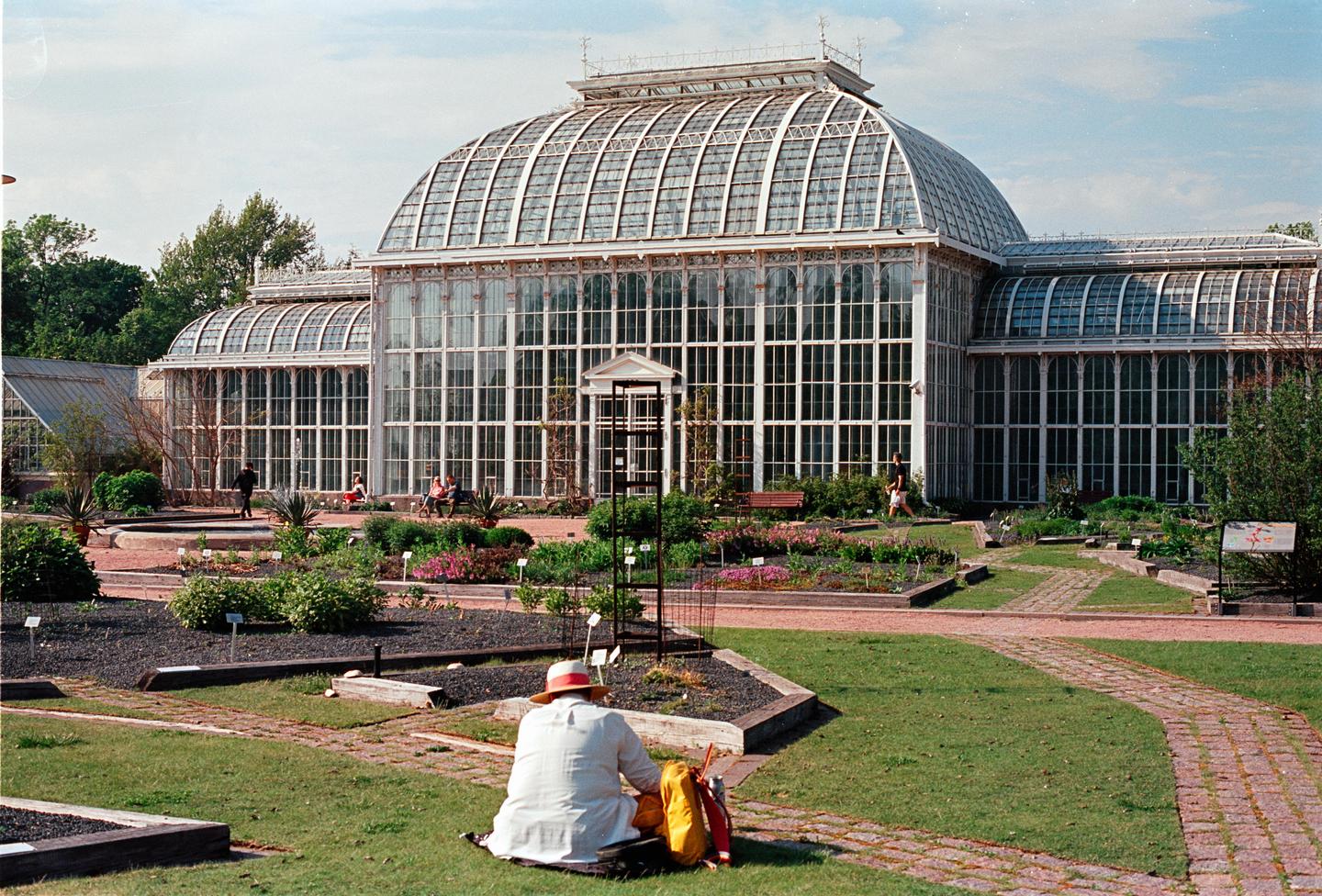 A person wearing a hat sits on the grass in front of a large glass conservatory with gardens and brick paths.