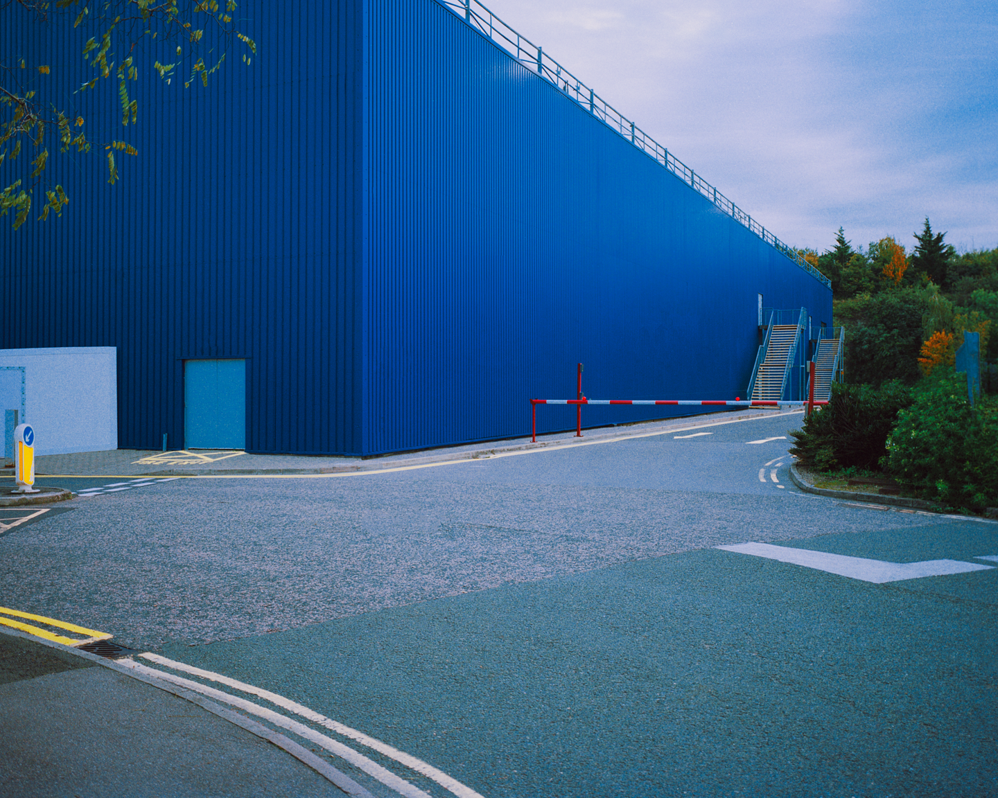 A large blue building with a barrier across a road and trees in the background is visible under a cloudy sky.