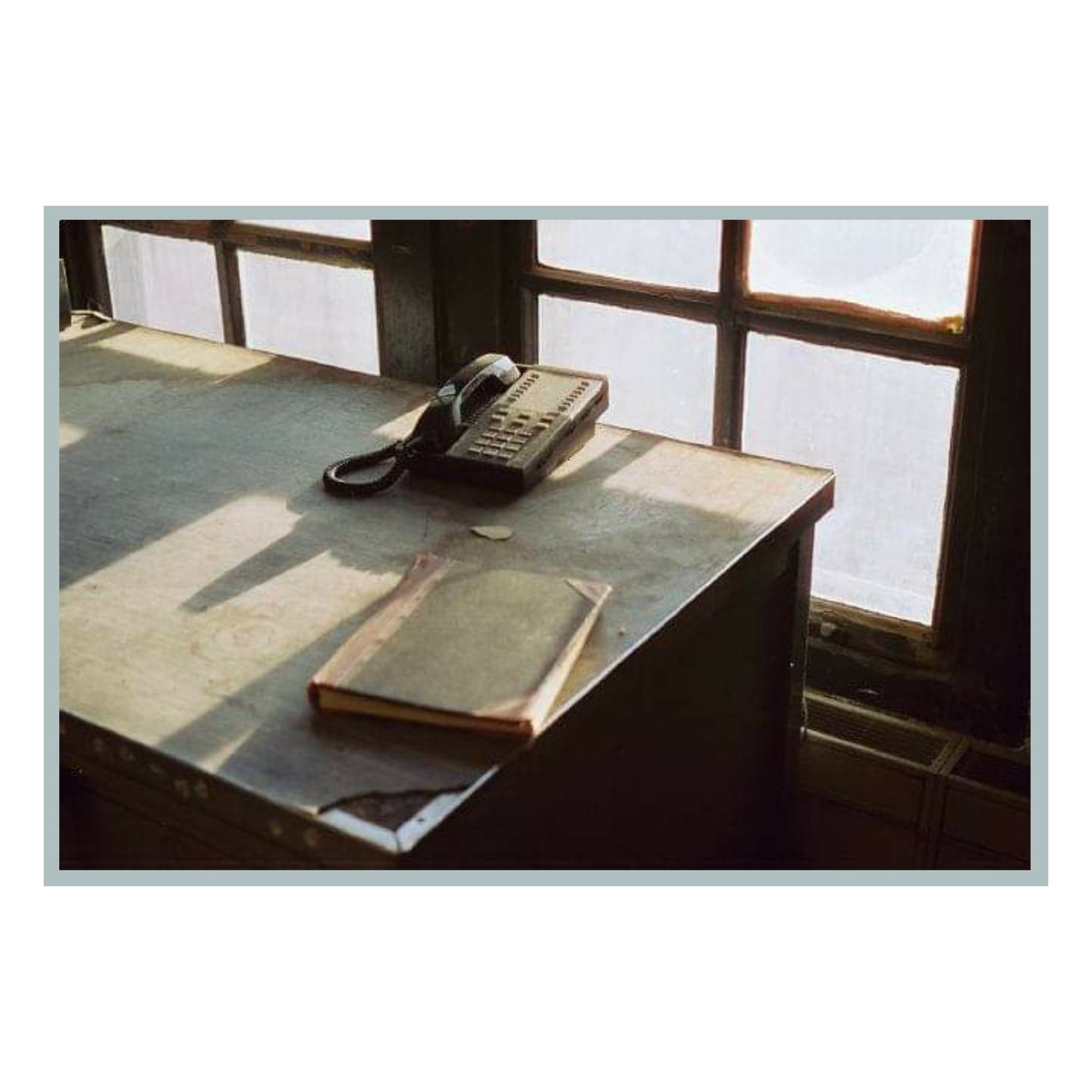 A telephone and a book sit on a desk next to a window with light streaming through.
