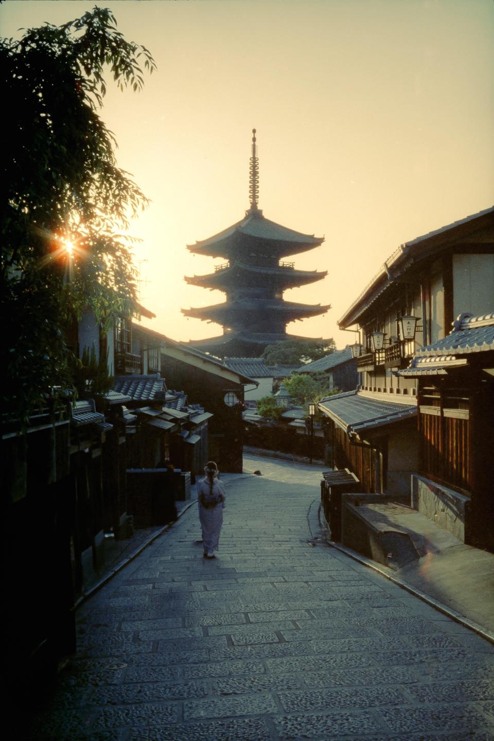 A woman walks down a cobblestone street towards a large pagoda at sunset in Kyoto.