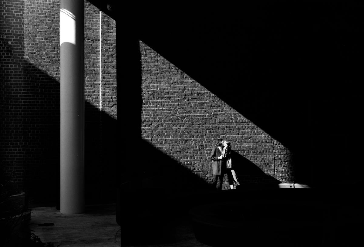 A couple stands in a patch of sunlight against a brick wall, with a large column to the left.