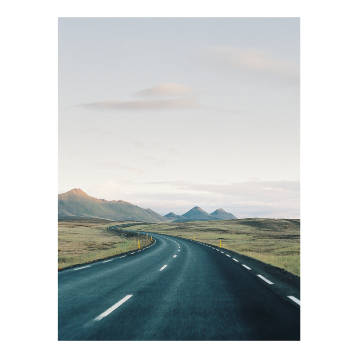 A winding road stretches through a vast, grassy landscape towards distant mountains under a pale sky.
