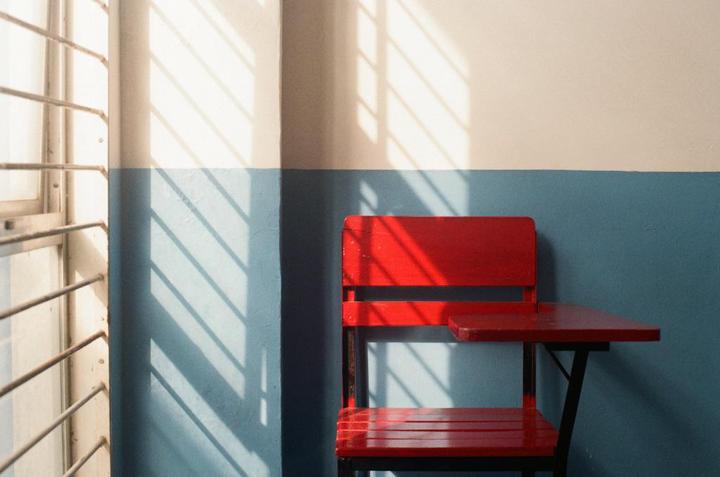 A red school desk sits against a two-toned wall with window shadows cast upon it.