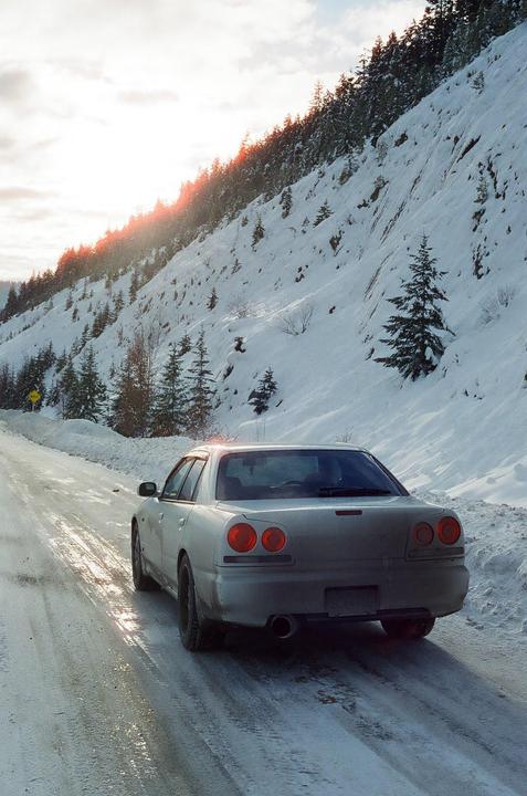 A silver car drives on a snowy road next to a snow-covered mountain with evergreen trees.