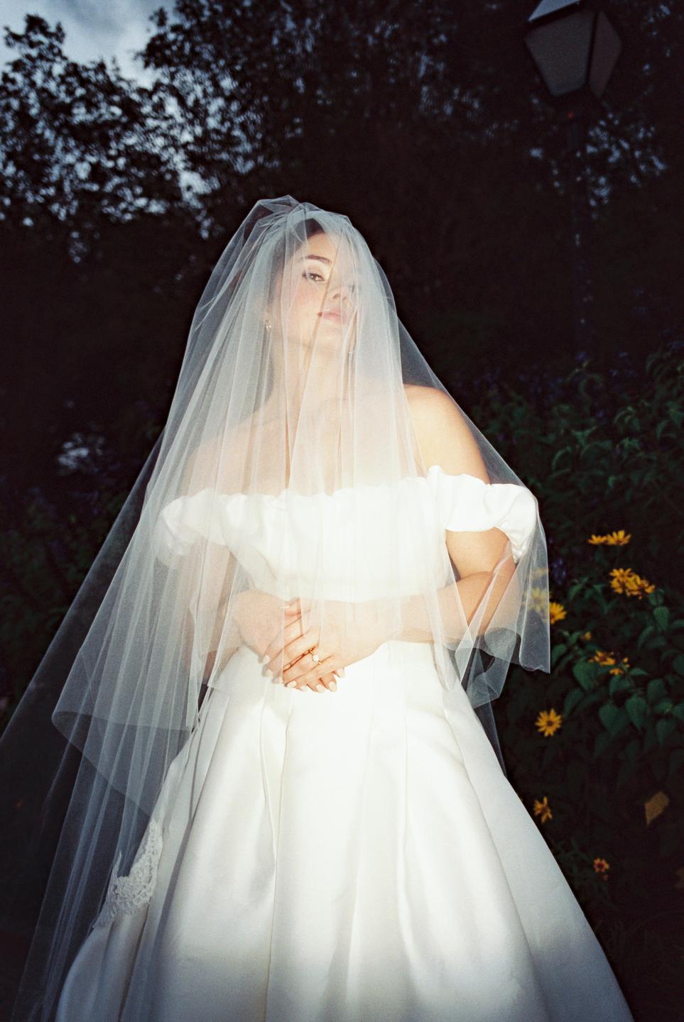 A woman in a white wedding dress and veil stands in a garden at dusk.