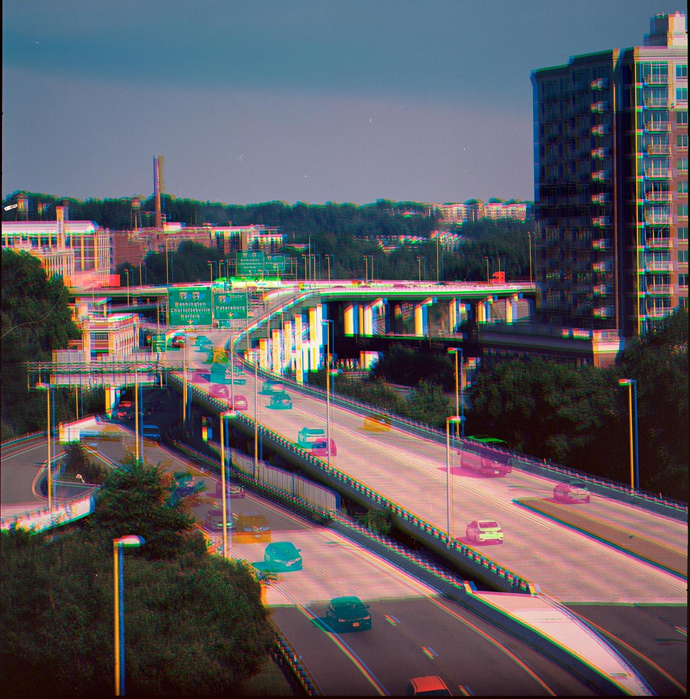 Cars drive on a multi-level highway system with buildings and trees in the background.