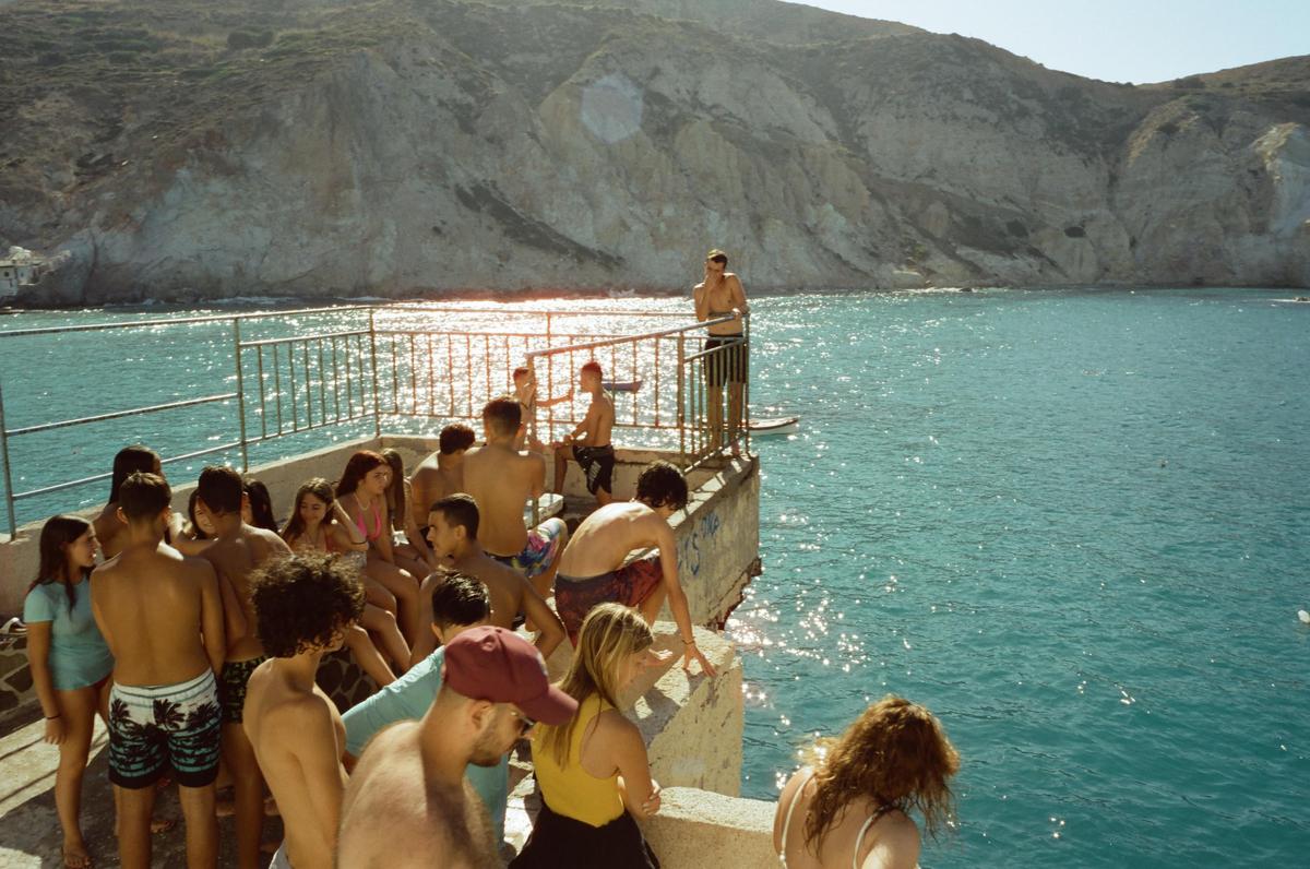 A group of young people gather on a concrete pier overlooking a bright blue sea with a rocky mountain in the background.