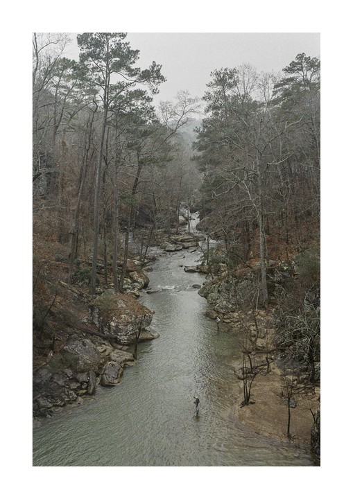 Two fishermen stand in a river surrounded by a forest with mostly bare trees under a hazy sky.