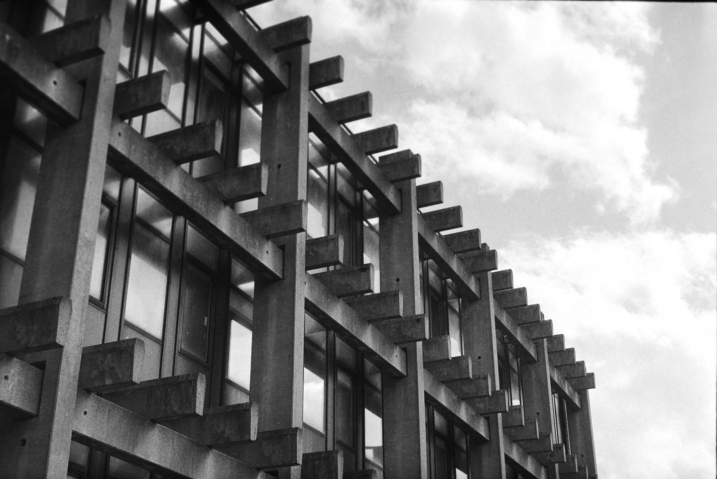 A brutalist building with many windows is shown from a low angle against a cloudy sky.