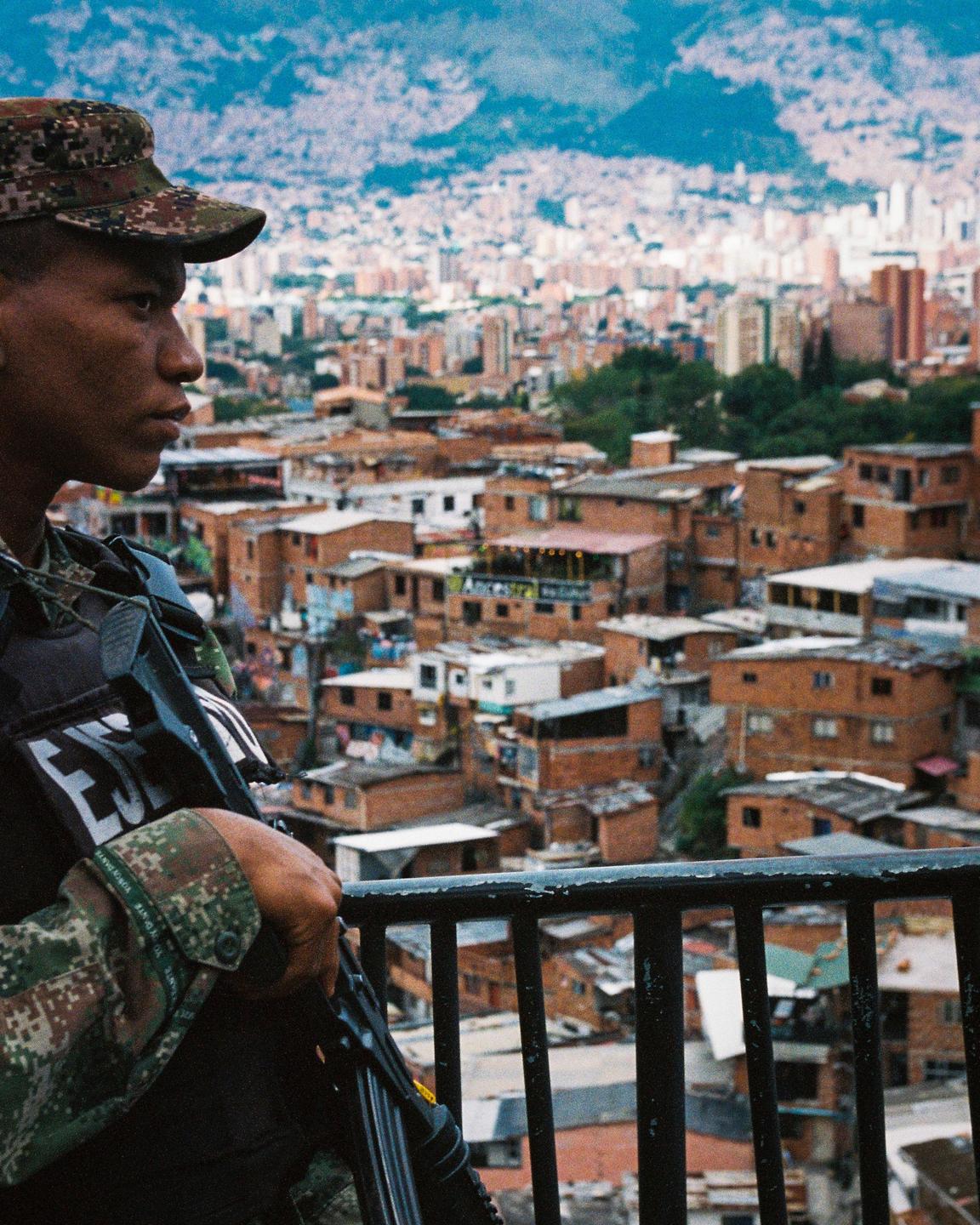 A man in military fatigues and a cap holds a rifle while looking out over a city built into a hillside.