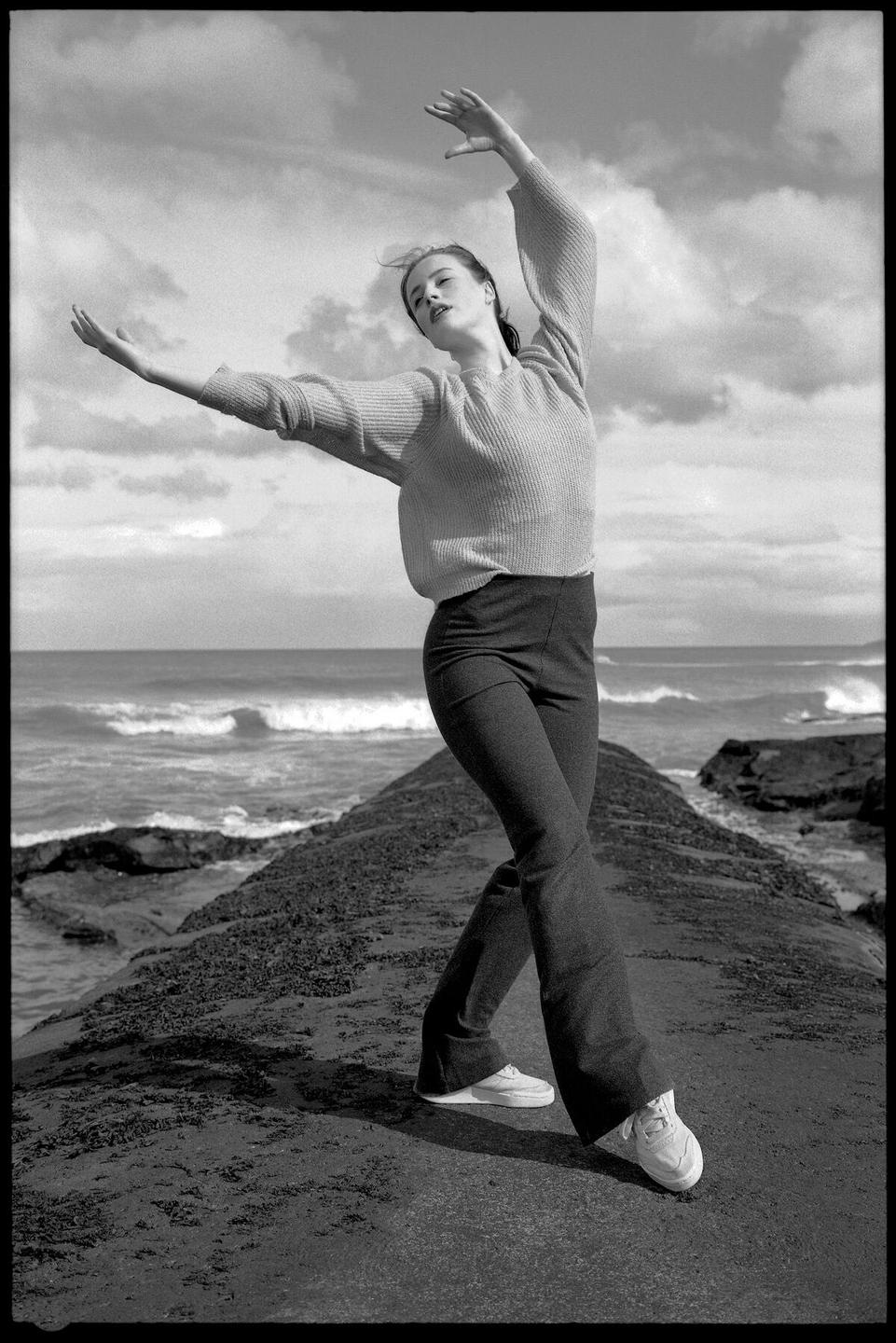 A woman in a sweater and pants poses with her arms raised on a rocky jetty by the sea.