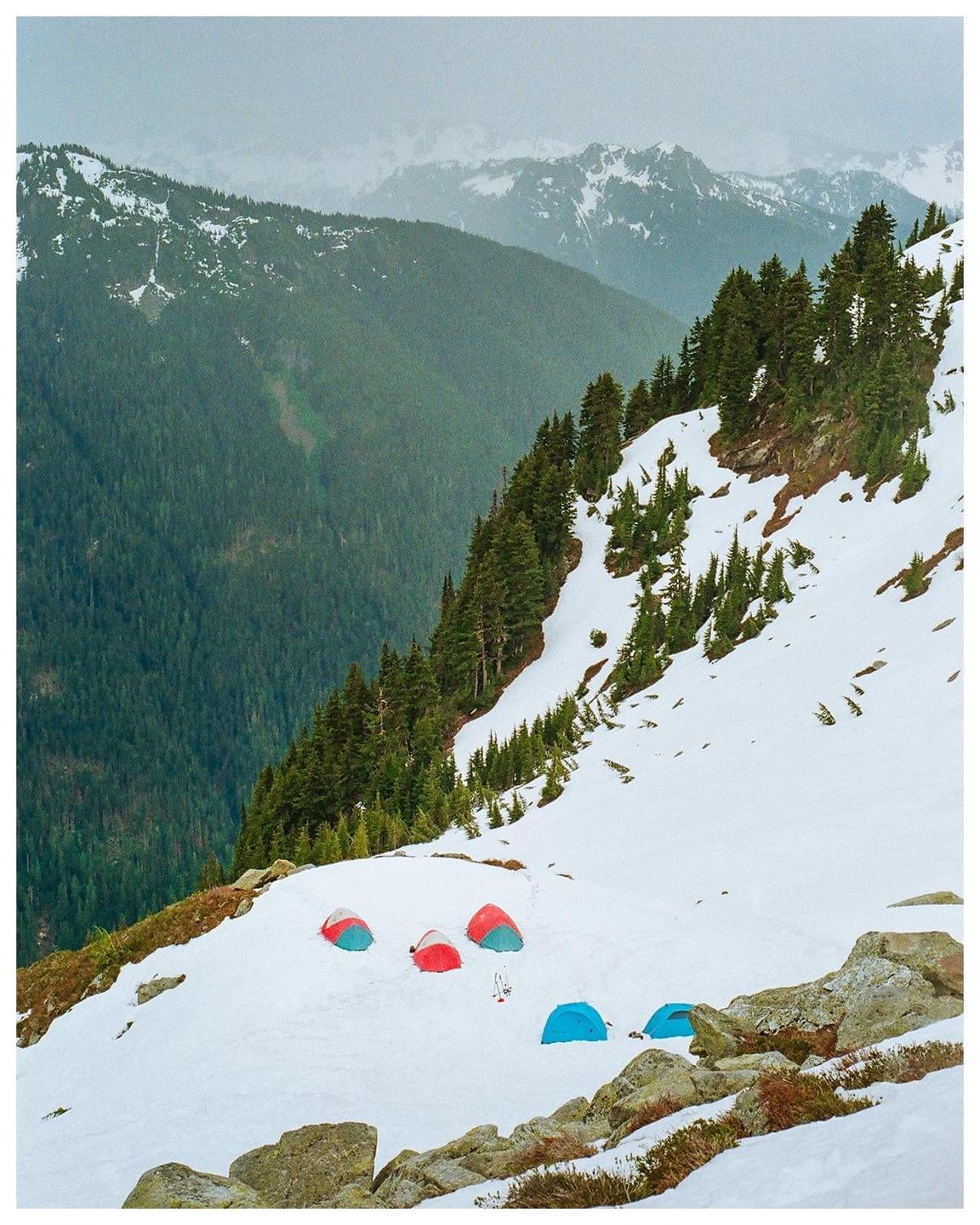 Several colorful tents are pitched on a snow-covered mountain slope with a forest and more mountains in the background.