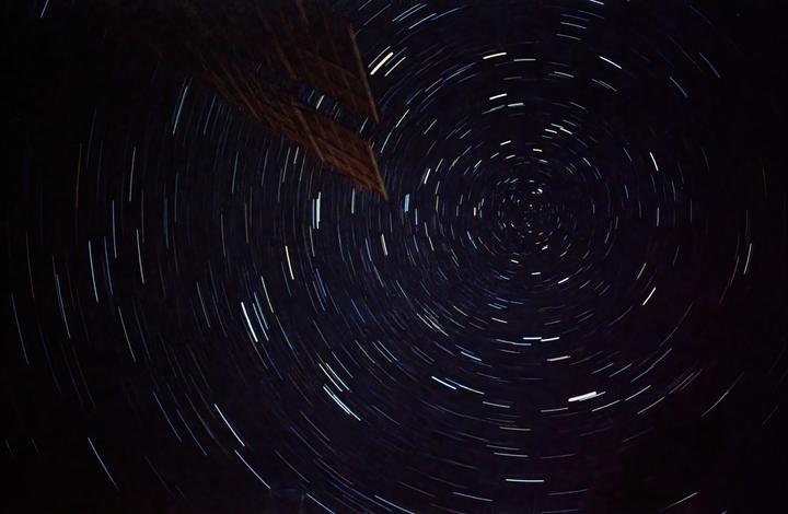 A long exposure photograph captures the circular star trails in the night sky, with a dark structure visible in the upper left corner.