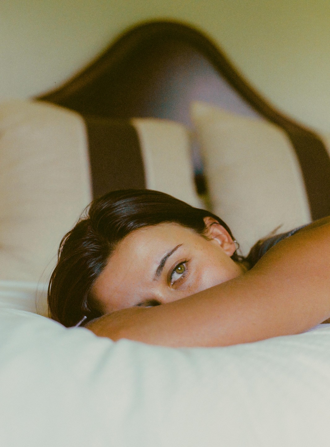A woman with dark hair and green eyes lies on a bed, resting her head on her arm.