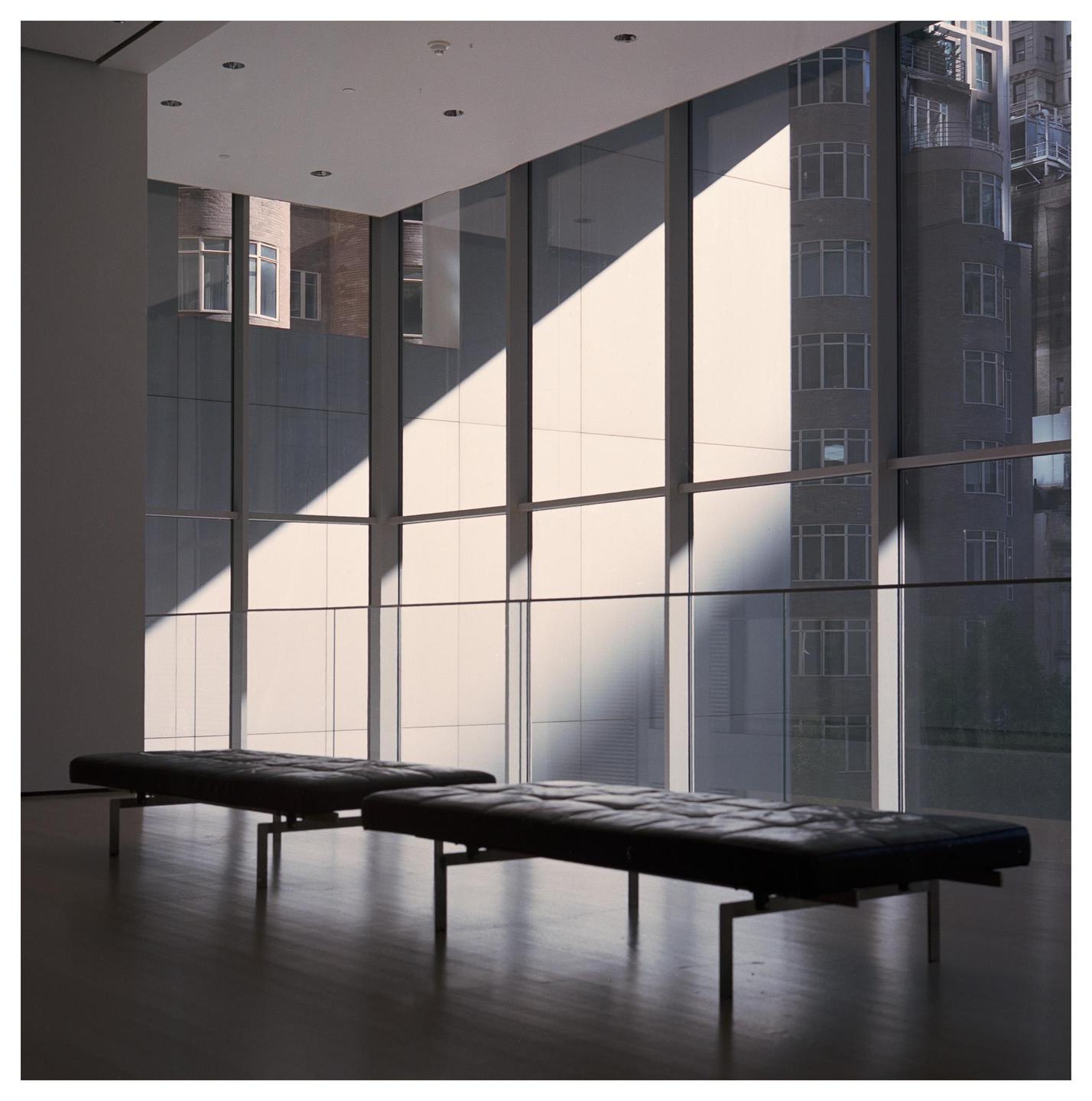 Two black benches sit on a dark wooden floor in a room with large windows looking out onto city buildings, with sunlight casting diagonal shadows across the windows.