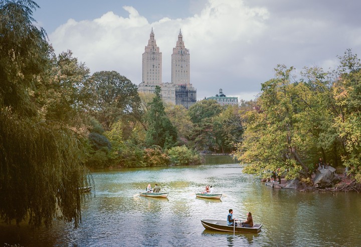 Several couples row boats on a lake in Central Park with tall buildings visible in the background.