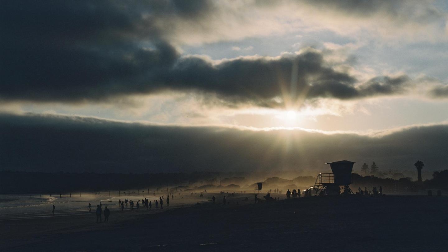 The sun shines through clouds over a beach with many people and a lifeguard stand.