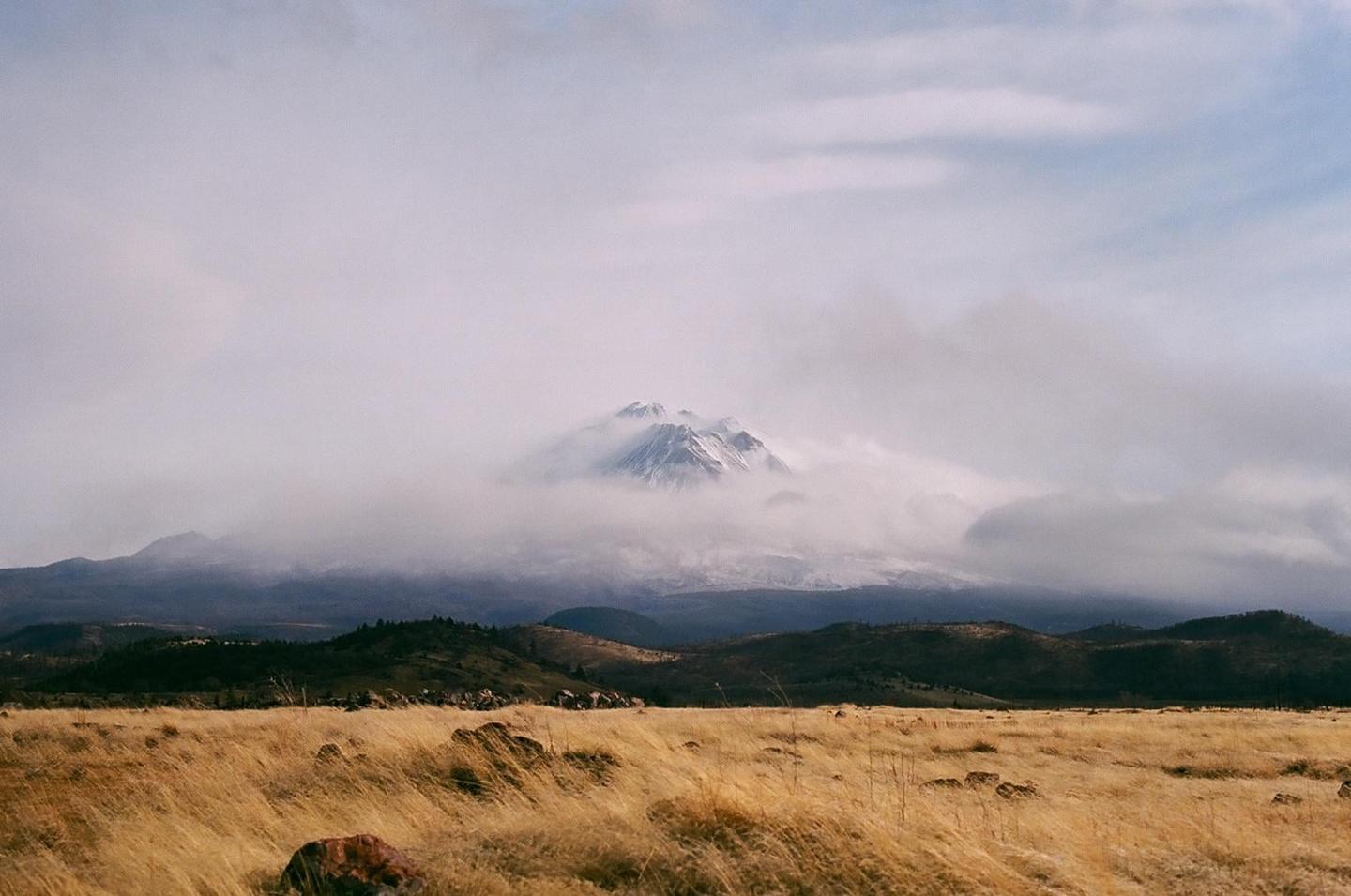 A snow-capped mountain peak is partially obscured by clouds above a field of golden grass and rolling hills.