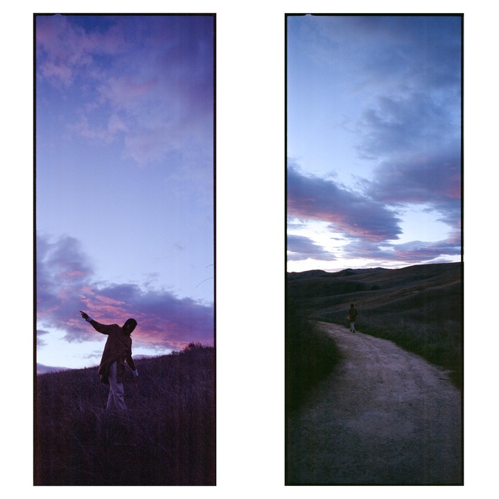 A person stands in tall grass on a hill under a purple and pink sunset sky, and another person walks along a dirt path through rolling hills under a similar sky.
