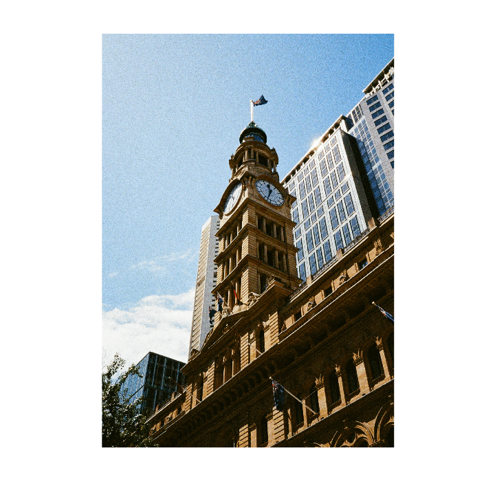 A tall clock tower with a flag on top stands against a bright blue sky, surrounded by modern buildings.