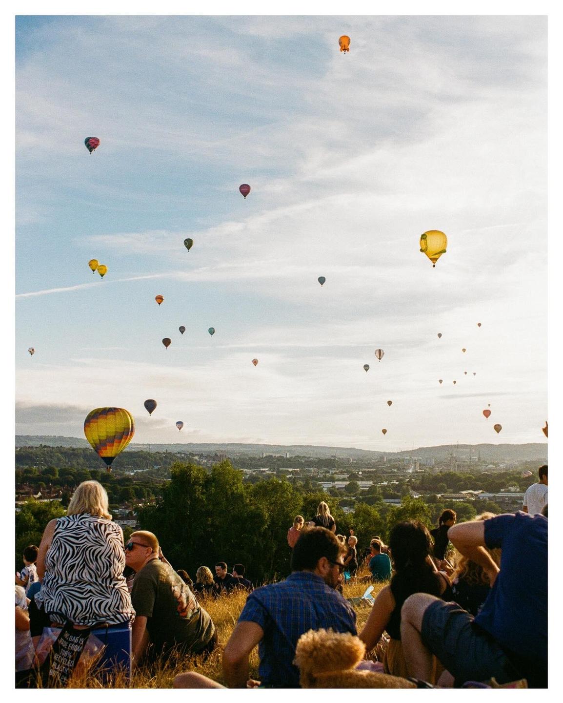 A crowd of people sit on a grassy hill watching hot air balloons float over a city.