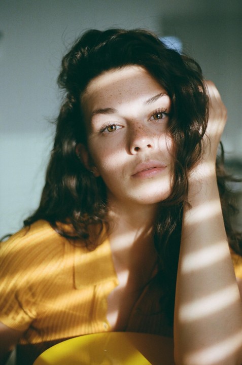 A woman with long, dark, curly hair and freckles looks directly at the viewer, with sunlight casting shadows across her face and body.