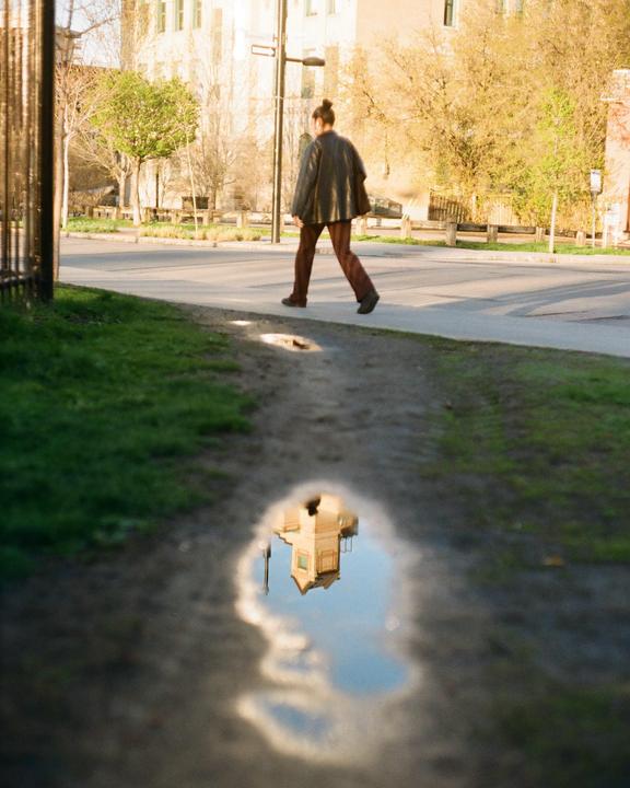 A person walks on a sidewalk next to a street with a reflection of a building and sky in a puddle in the foreground.