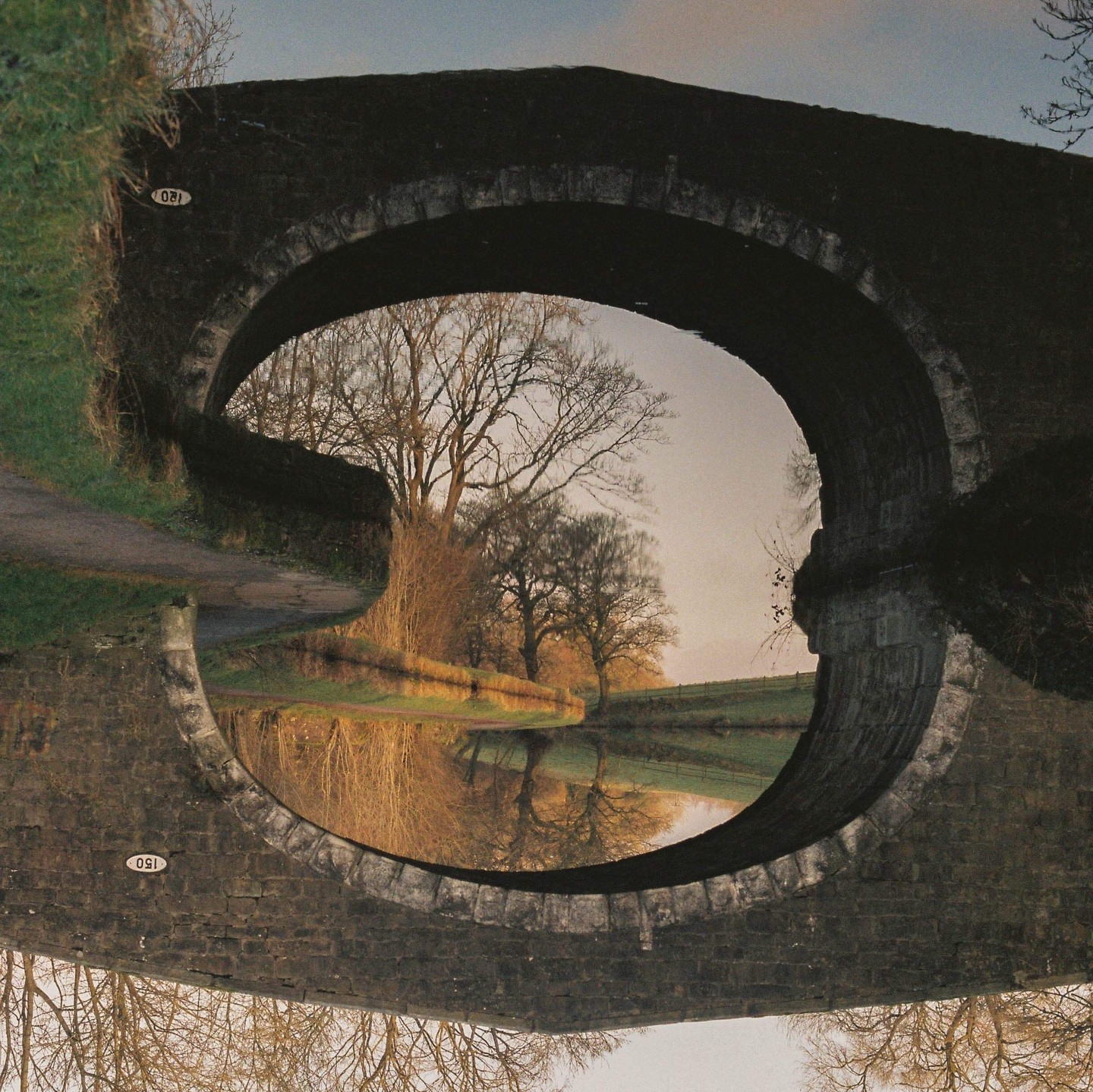 A canal and trees are visible through the arch of a stone bridge, with the scene reflected in the water.