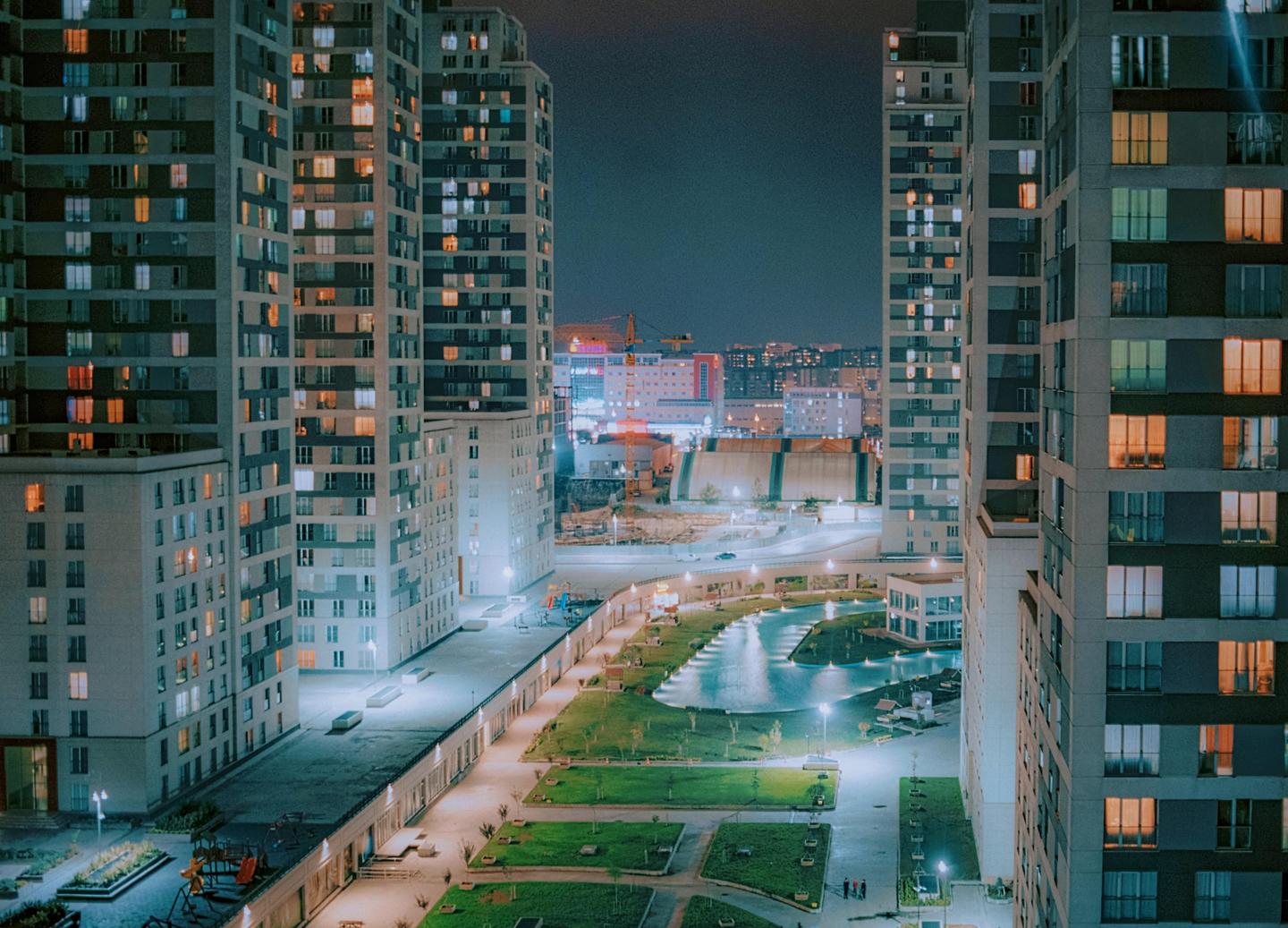 Tall apartment buildings with many illuminated windows flank a park with a winding water feature at night.