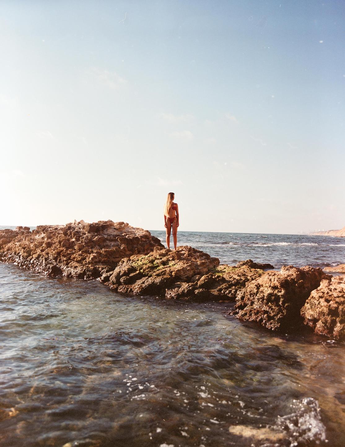 A woman stands on a rocky outcrop in the ocean, looking out at the water.