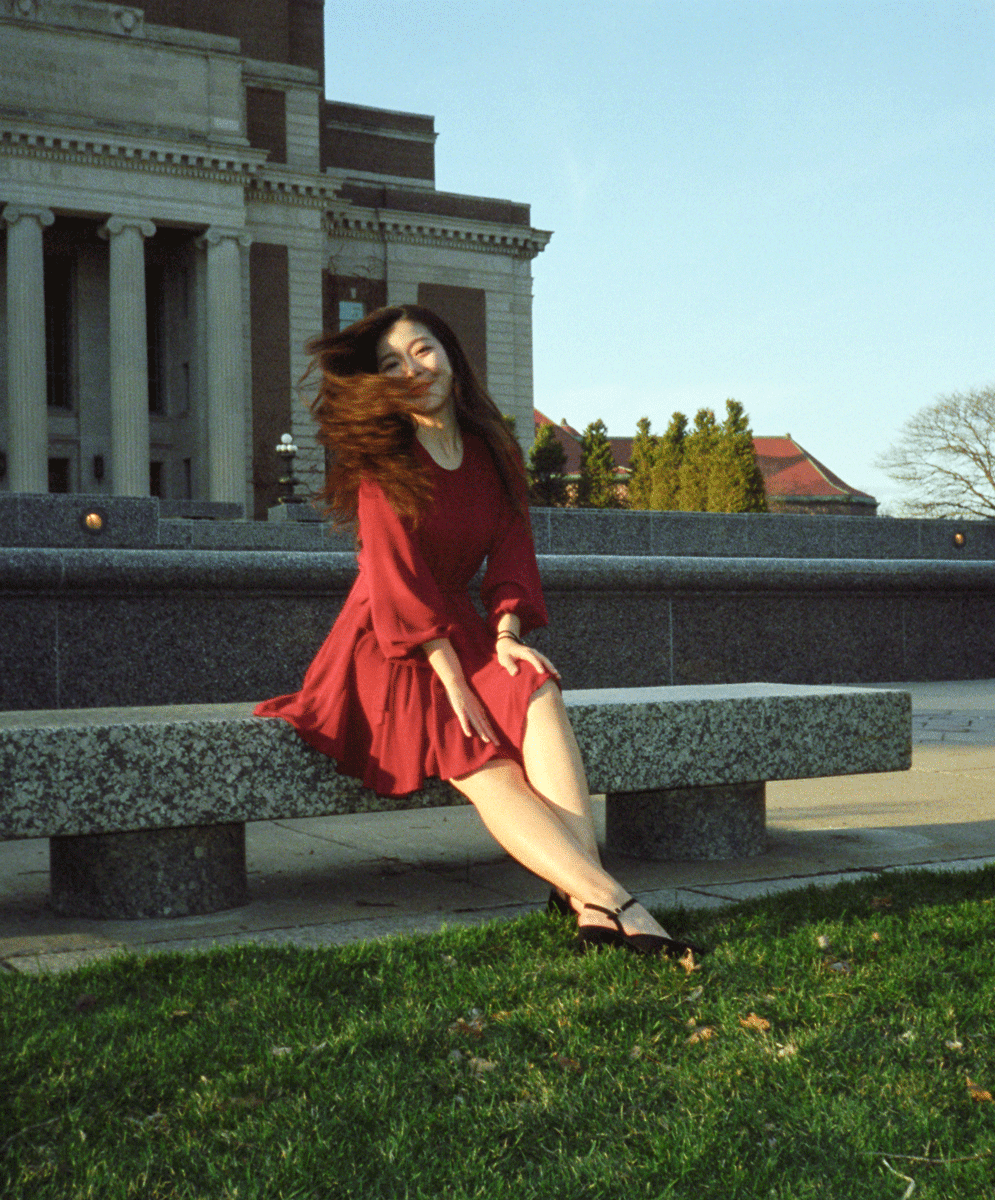 A woman in a red dress sits on a stone bench with a large building in the background.