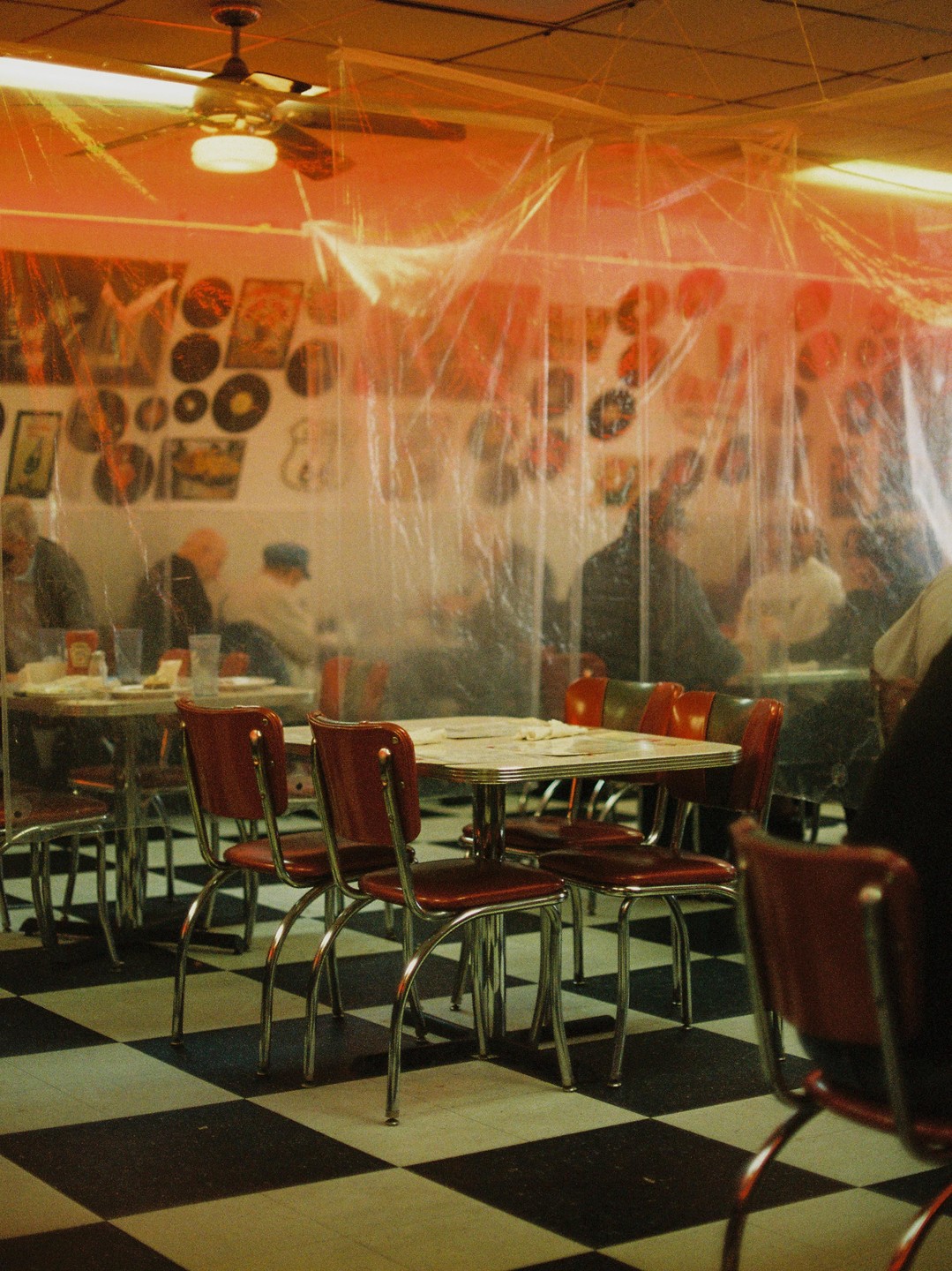 A diner with red chairs and checkered floor has plastic sheeting hanging from the ceiling, separating tables where people are seated.
