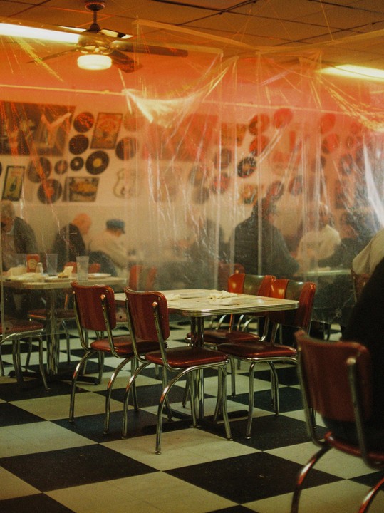 A diner with red chairs and checkered floor has plastic sheeting hanging from the ceiling, separating tables where people are seated.